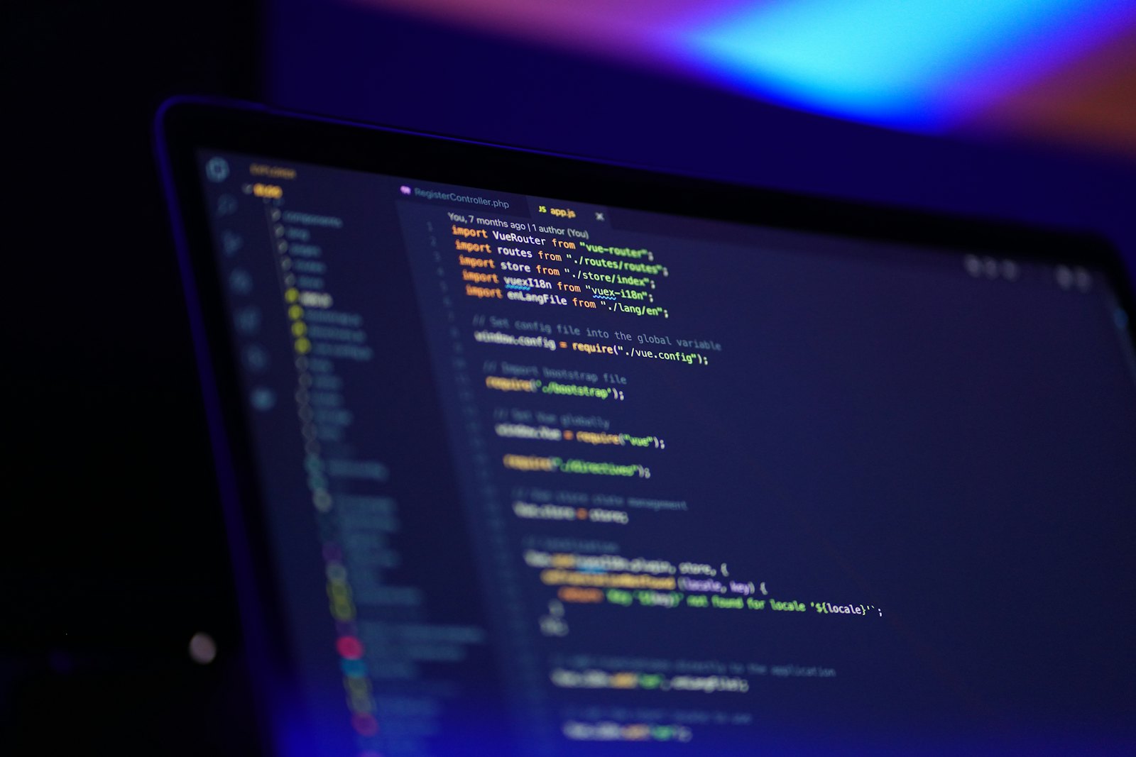 A laptop screen displaying colorful source code in a dark room lit by blue and purple light